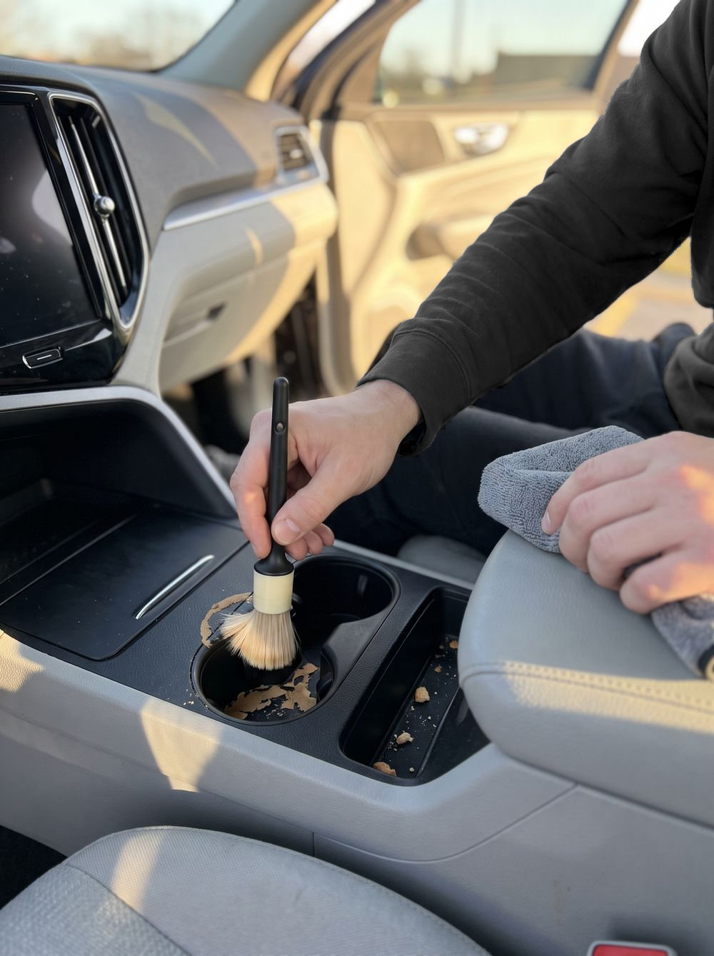 A soft detailing brush lifts dried coffee from a cupholder in an SUV console, a gray cloth in the other hand, low sun through the open door.